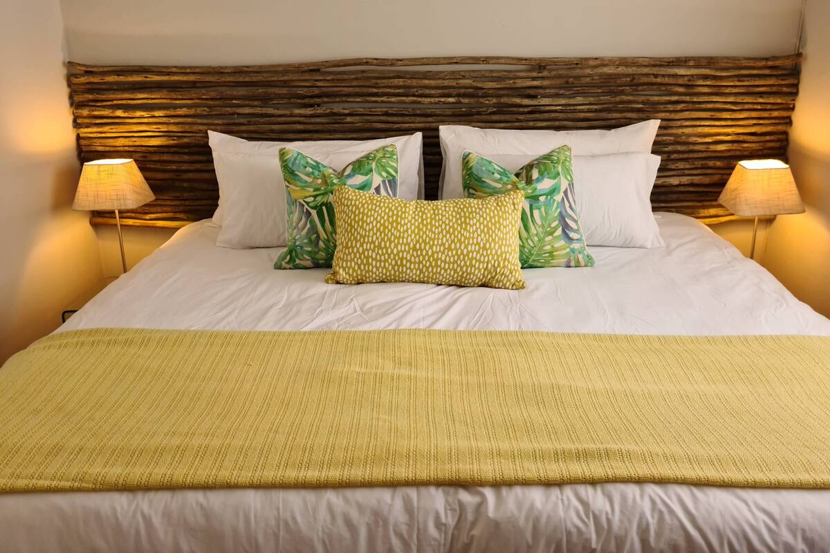 A neatly made bed with white linens and a yellow throw blanket, flanked by two lamps and a wooden headboard.