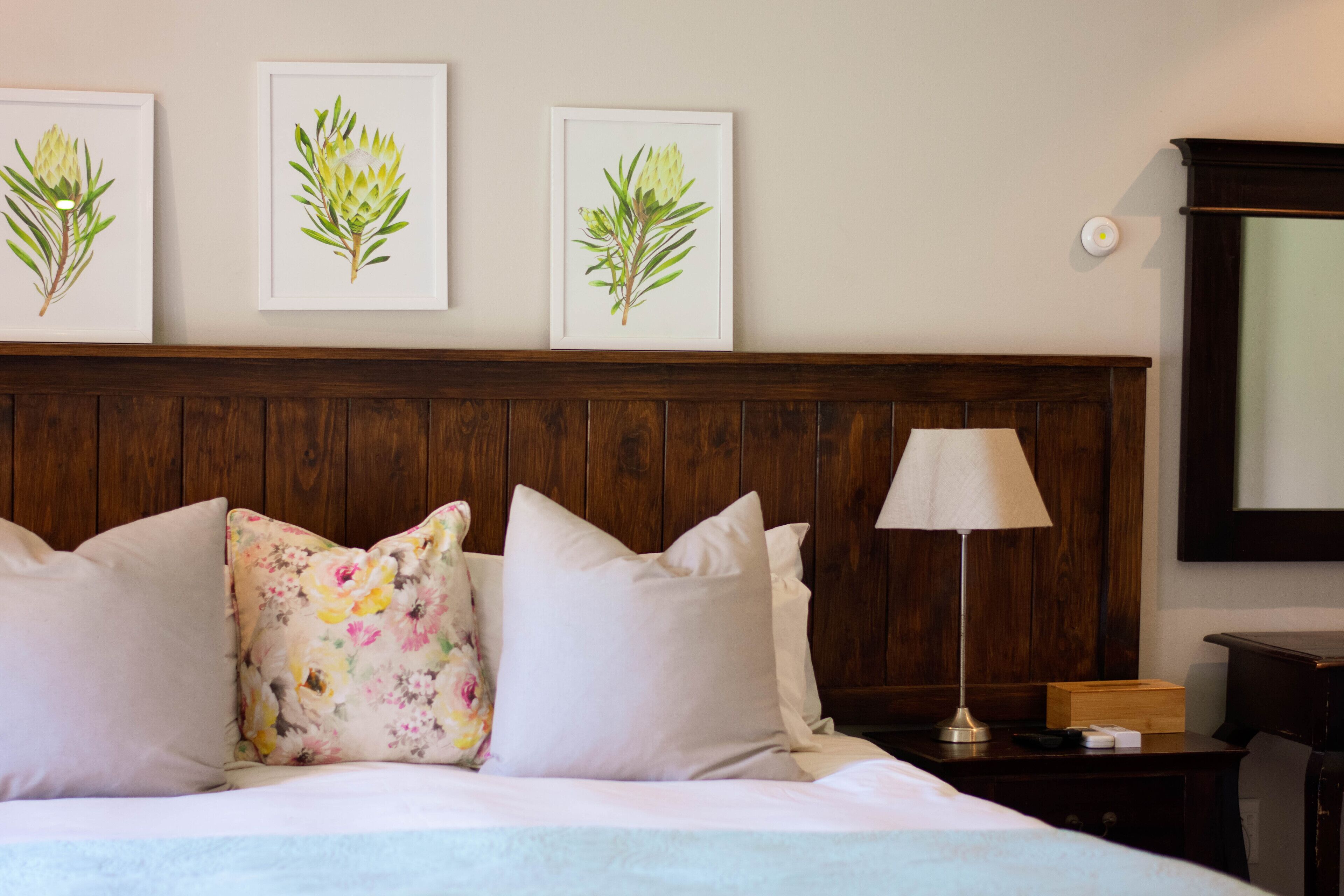 Ein ordentlich bezogenes Bett mit floralen Kissen, ein Nachttisch mit Lampe und gerahmte Pflanzenbilder an der Wand.