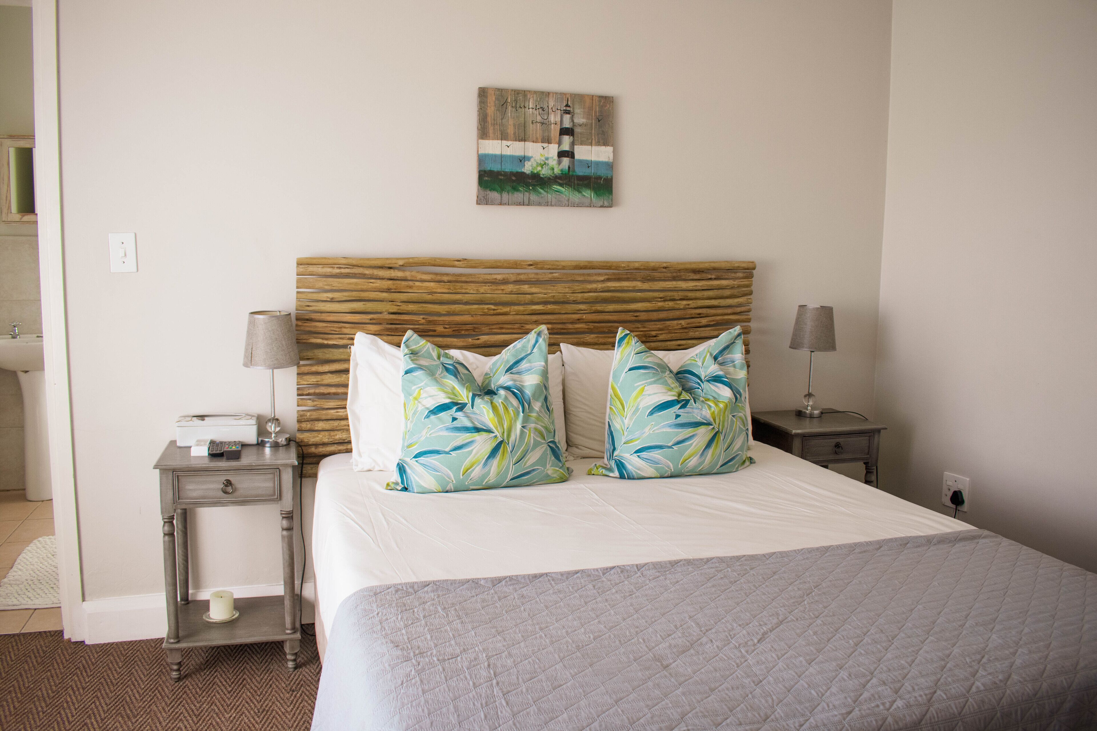 A neatly made bed with a quilted cover, two matching pillows, a wooden headboard, a bedside table with a lamp, and a wall-mounted artwork depicting a lighthouse.