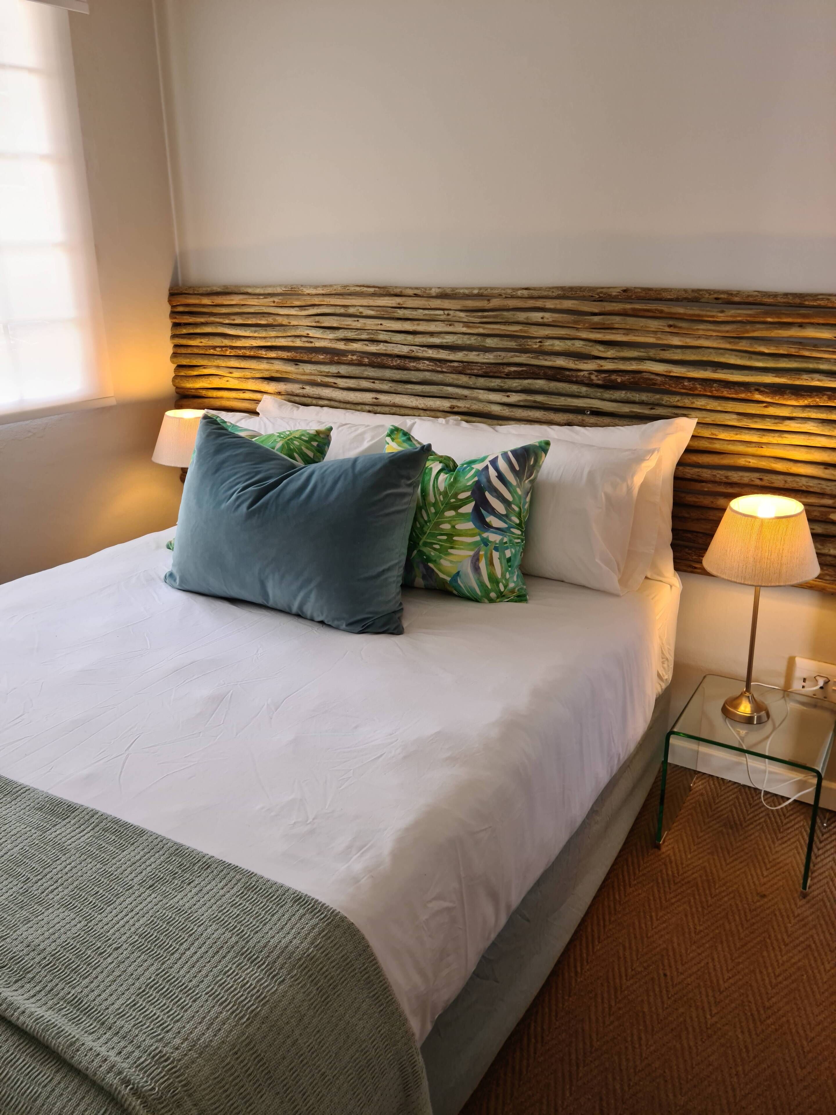 A neatly made bed with white linens and a green throw blanket, a wooden headboard, and a bedside lamp on a glass table.