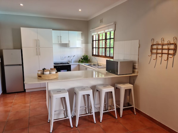 A modern kitchen with white cabinets, a built-in oven, a microwave, and a sink. There are three white bar stools and a small island with a sink.