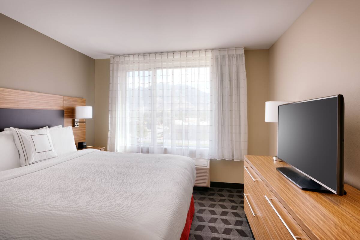 Ein Hotelzimmer mit einem großen Bett, einem Flachbildfernseher, einem Nachttisch mit Lampe und einem Fenster mit durchsichtigen Vorhängen.