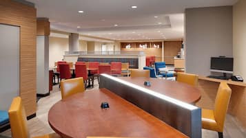 A modern hotel lobby with a central round table, multiple seating areas with red and yellow chairs, a kitchen area in the background, and a flat-screen TV on a desk.