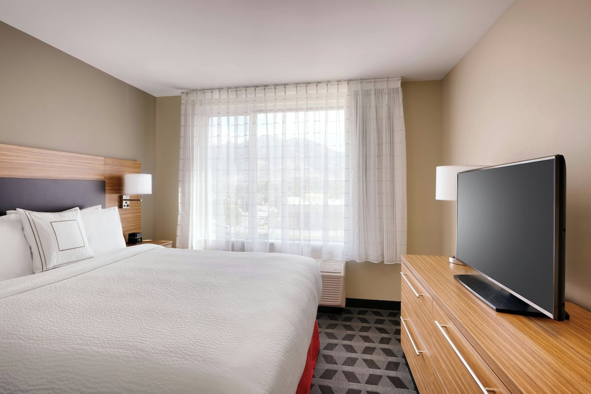Ein Hotelzimmer mit einem großen Bett, einem Flachbildfernseher, einer Holzkommode und einem Fenster mit durchsichtigen Vorhängen.