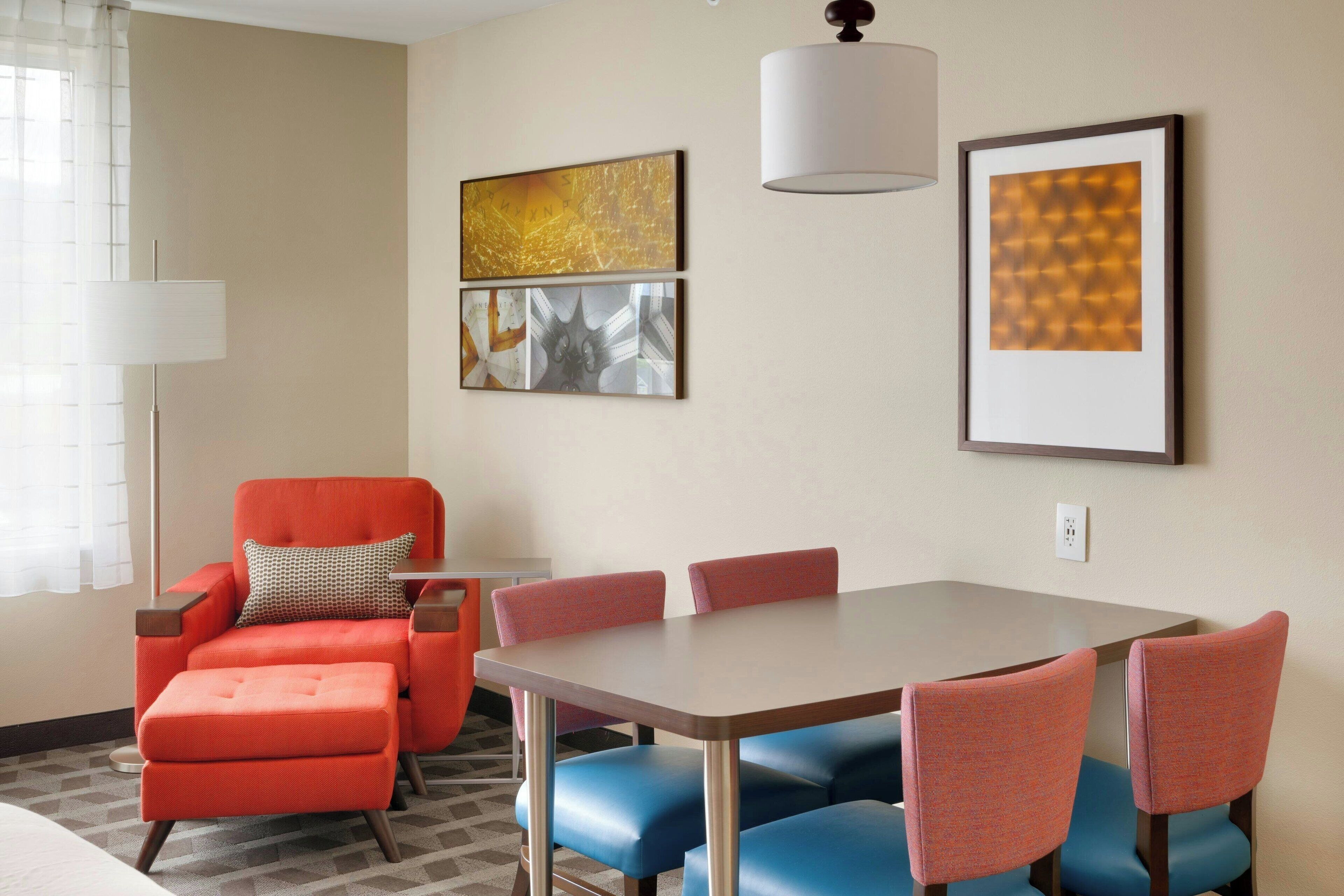 A hotel room with a dining table, four chairs, a red armchair, and two wall-mounted artworks.
