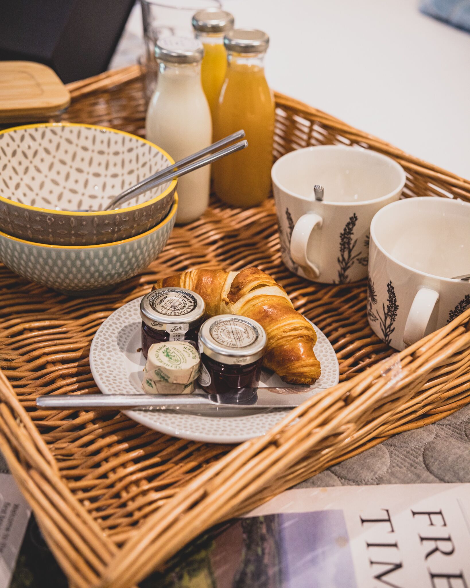 A breakfast tray with croissants, jam, honey, and coffee cups on a wicker table.
