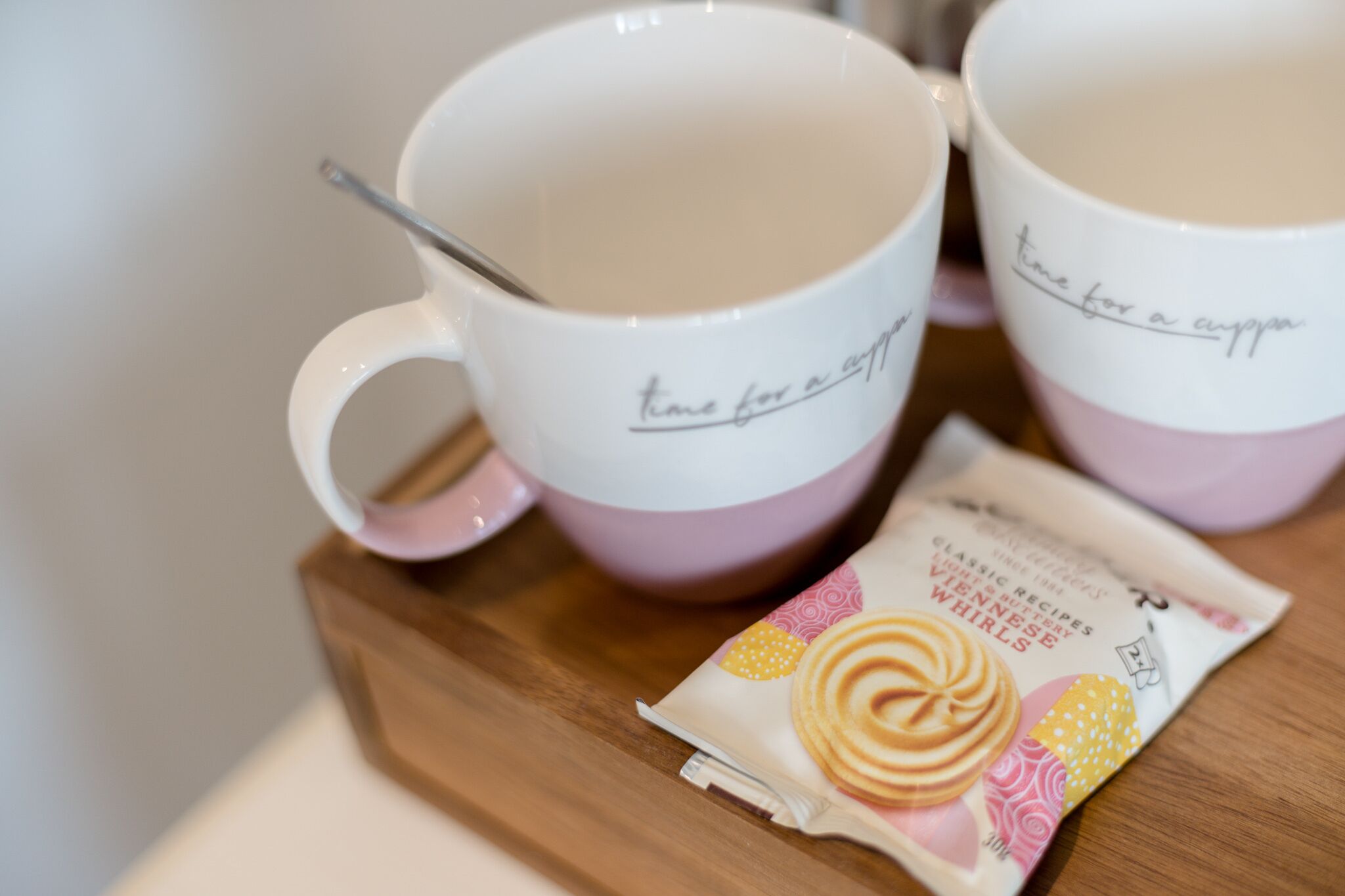 Dos tazas blancas y rosadas con una cuchara, un paquete de galletas y una bandeja de madera.