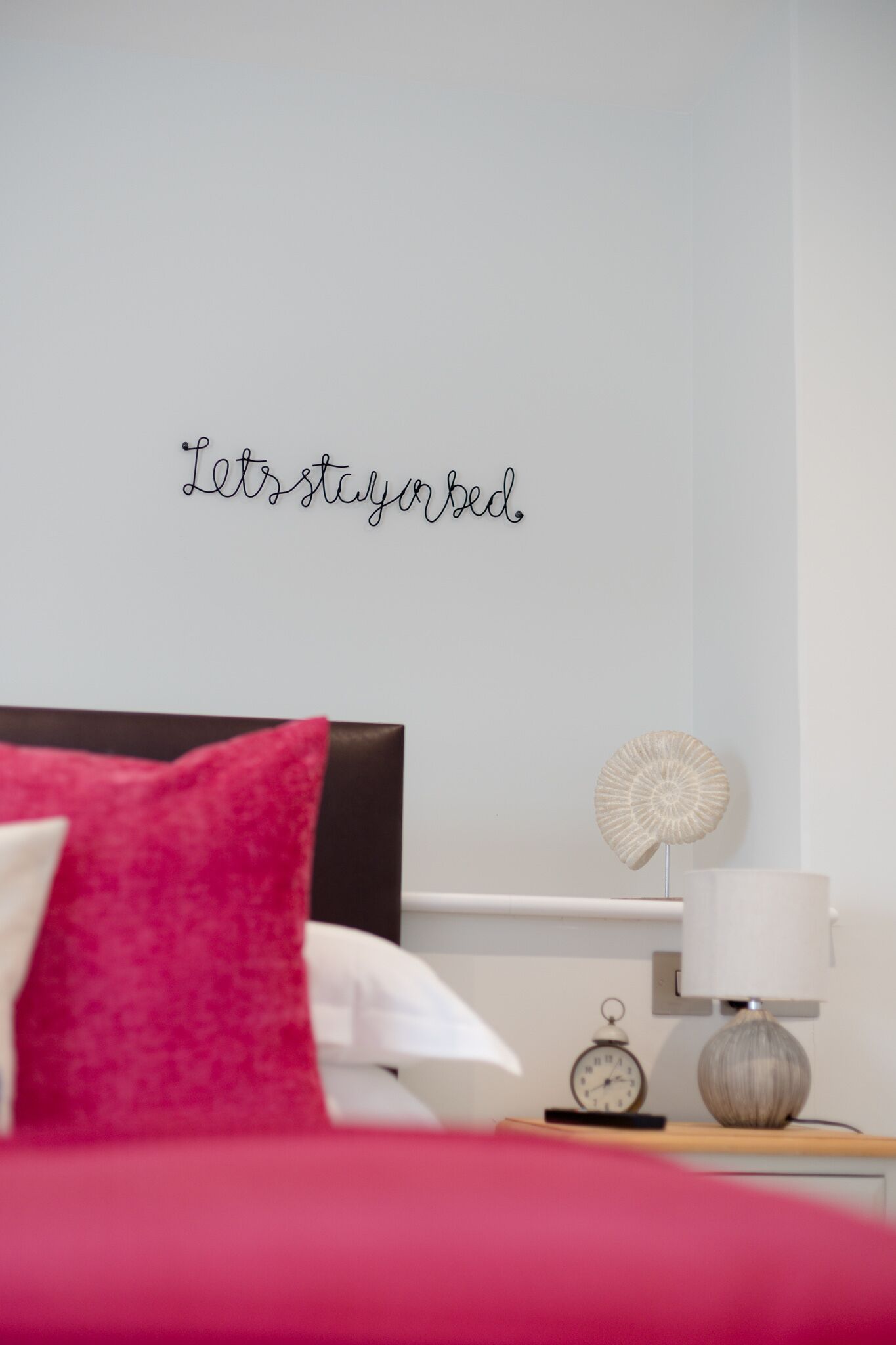 A neatly made bed with a red and white pillow, a wall clock, and a decorative wall piece that reads "Let's stay in bed."