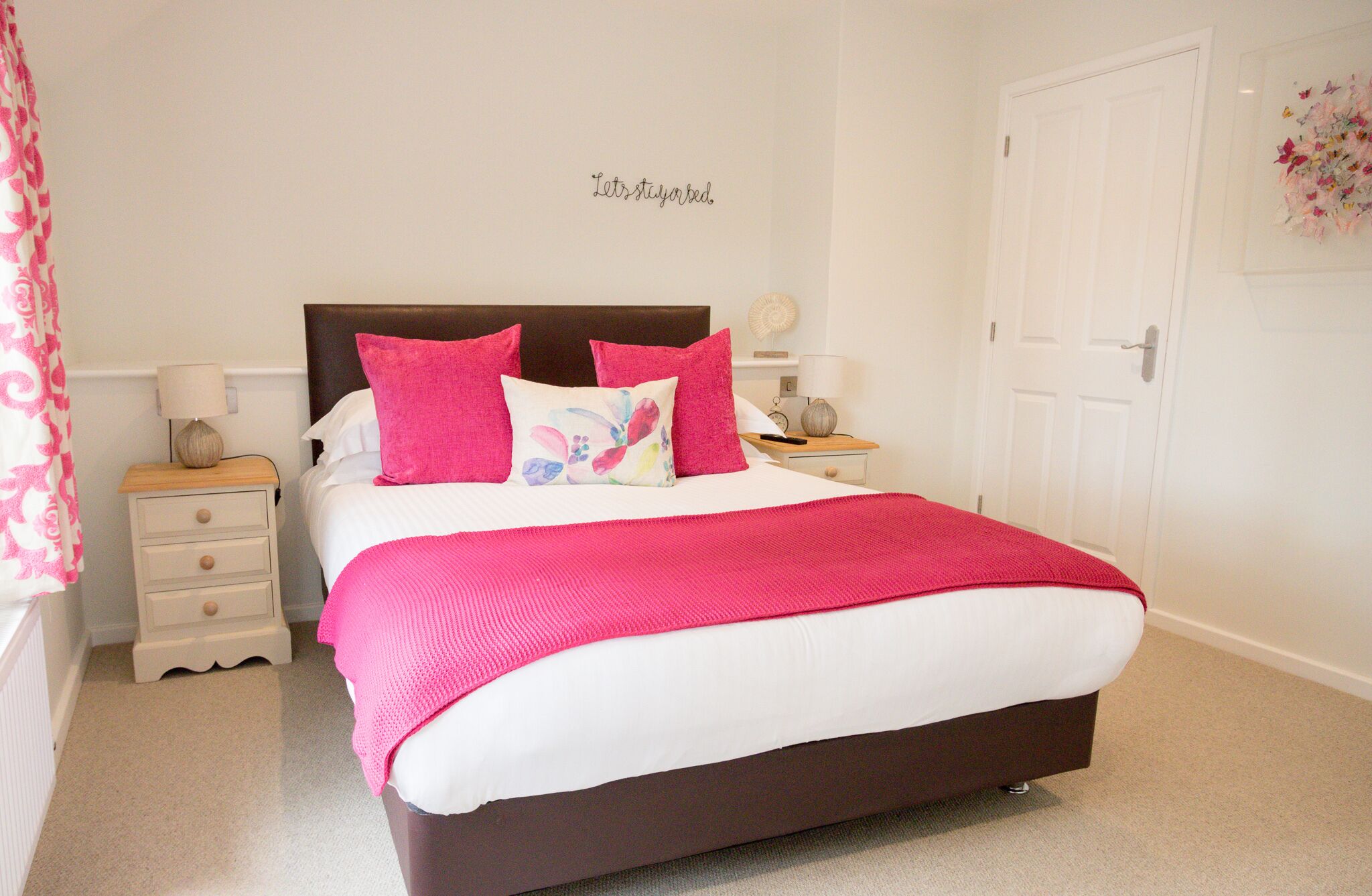 A neatly made bed with a white and pink color scheme, a headboard, and a bedside table with a lamp.