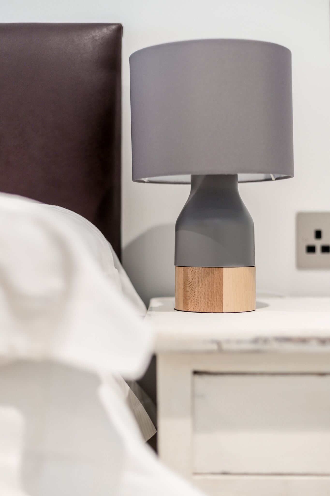 A modern bedside table with a lamp, a dark brown headboard, and a white bedside table.