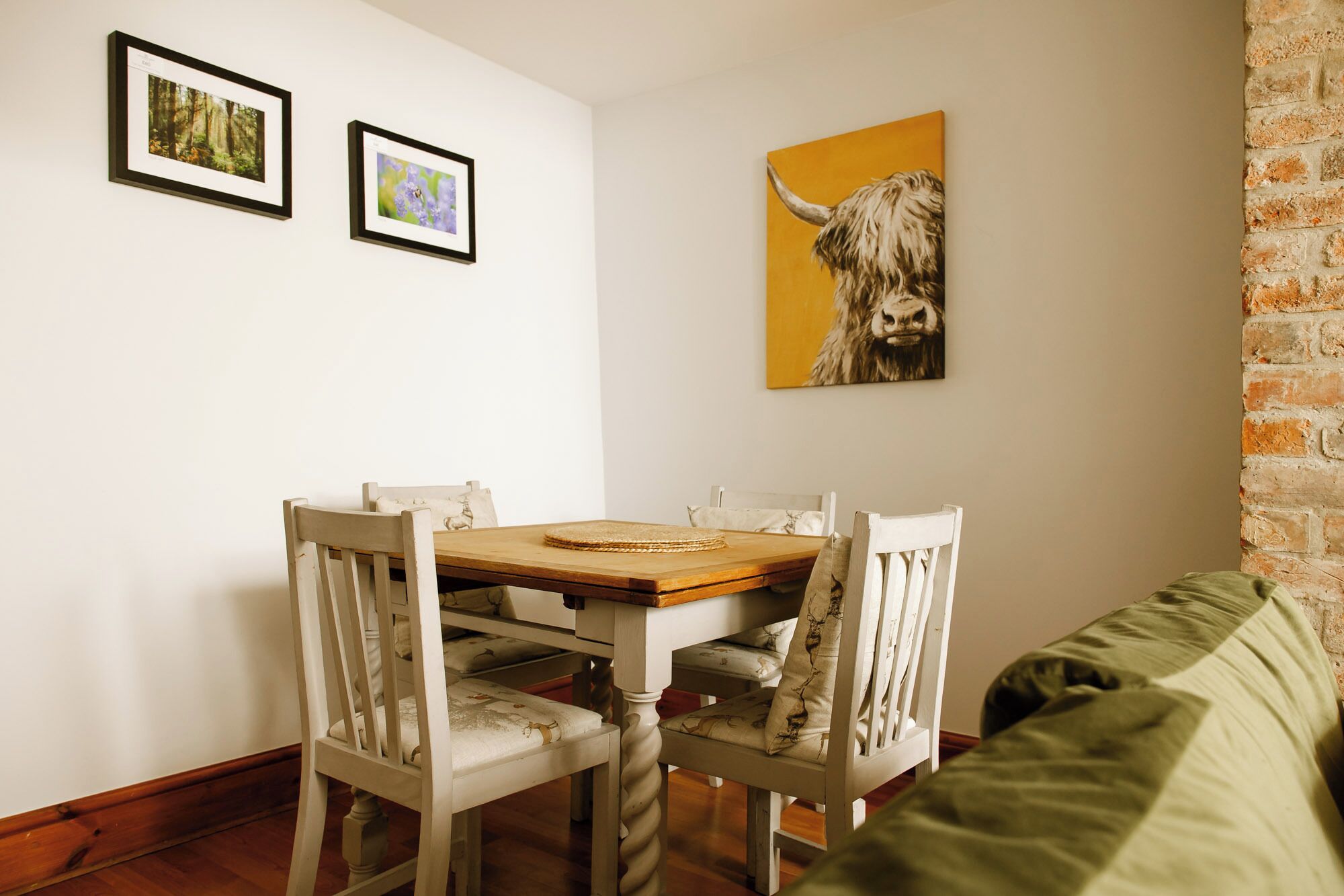 Uma sala de jantar com uma mesa de madeira e cadeiras brancas, um sofá verde e duas fotografias emolduradas na parede.