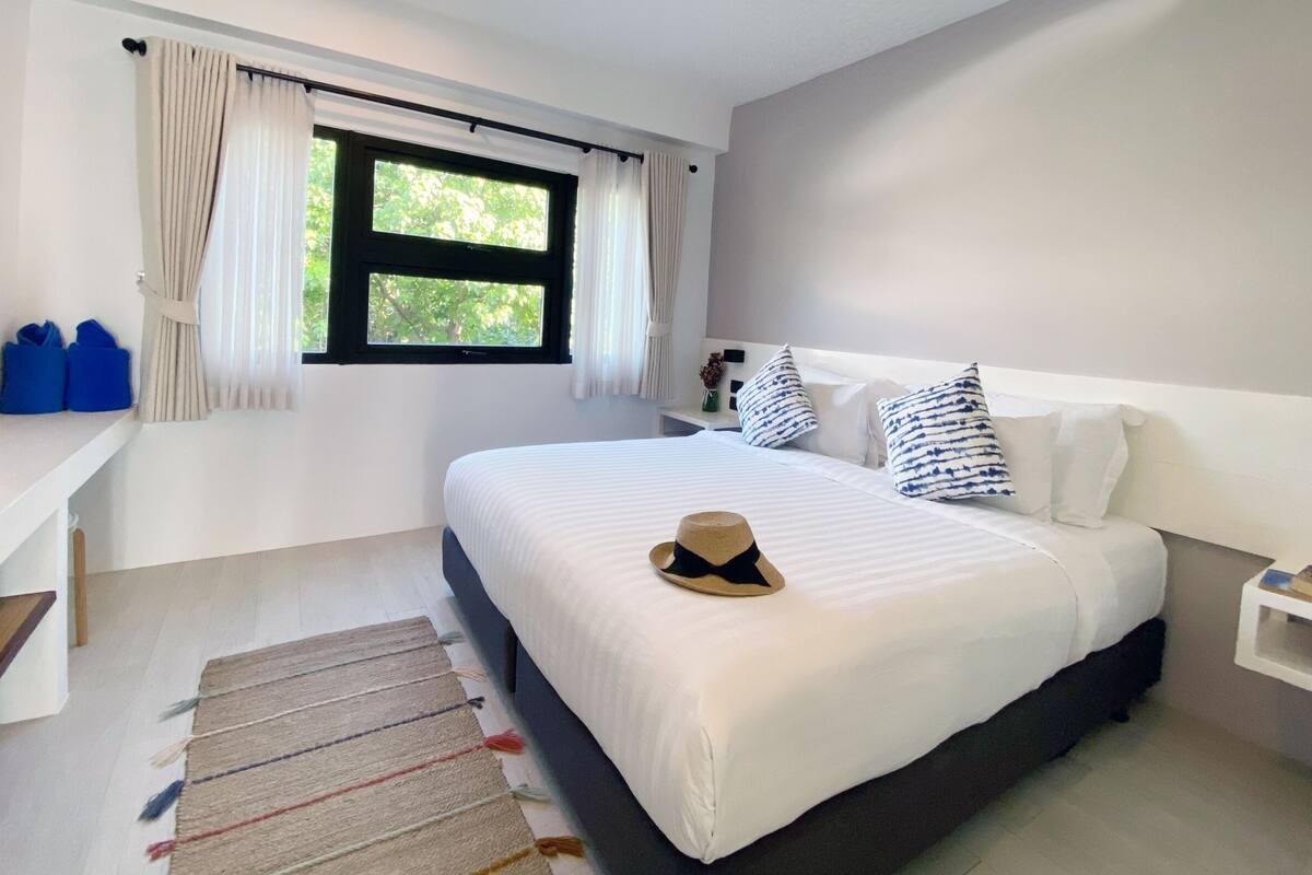 A bedroom with a large bed, a window with curtains, a bedside table with a vase, and a small shelf with books.