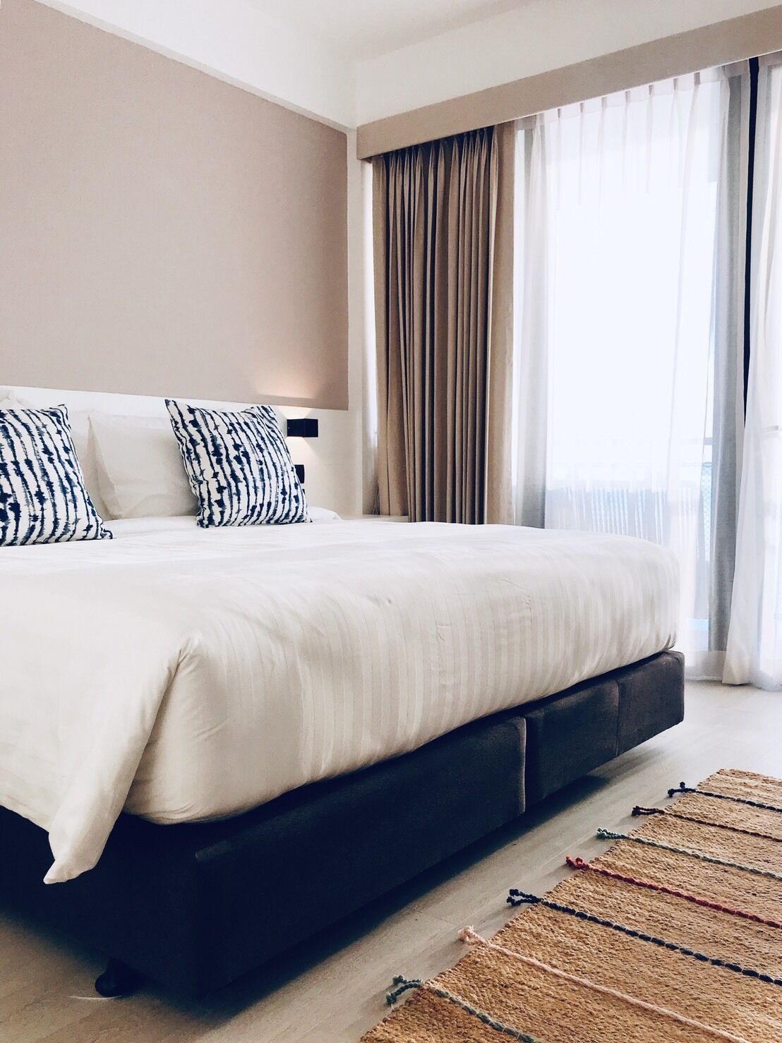 Una habitación de hotel moderna con una cama grande, un cabecero oscuro y vistas a través de cortinas de gasa.
