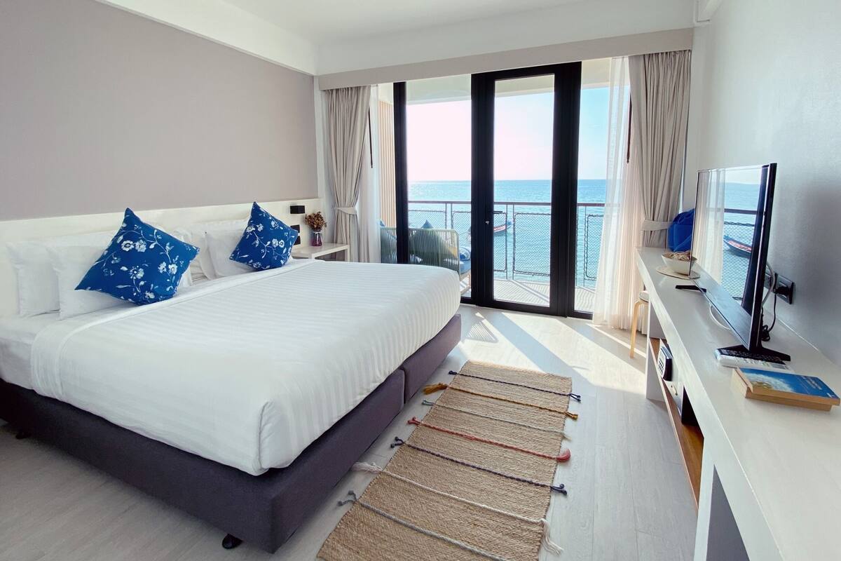 A hotel room with a large bed, a desk with a computer, and a view of the sea through a sliding door.