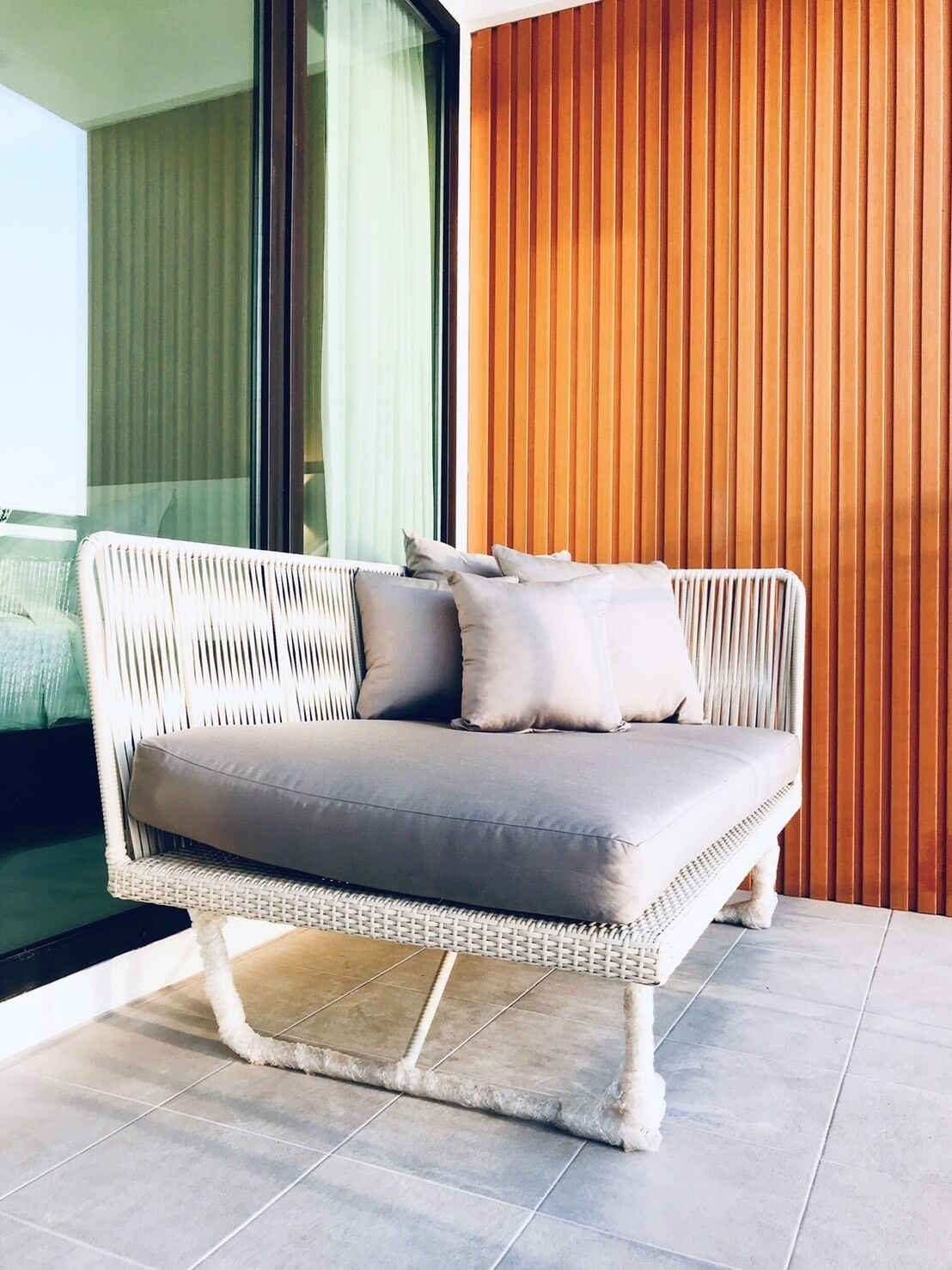 A wicker sofa with cushions, positioned on a tiled floor, adjacent to a window with vertical blinds.
