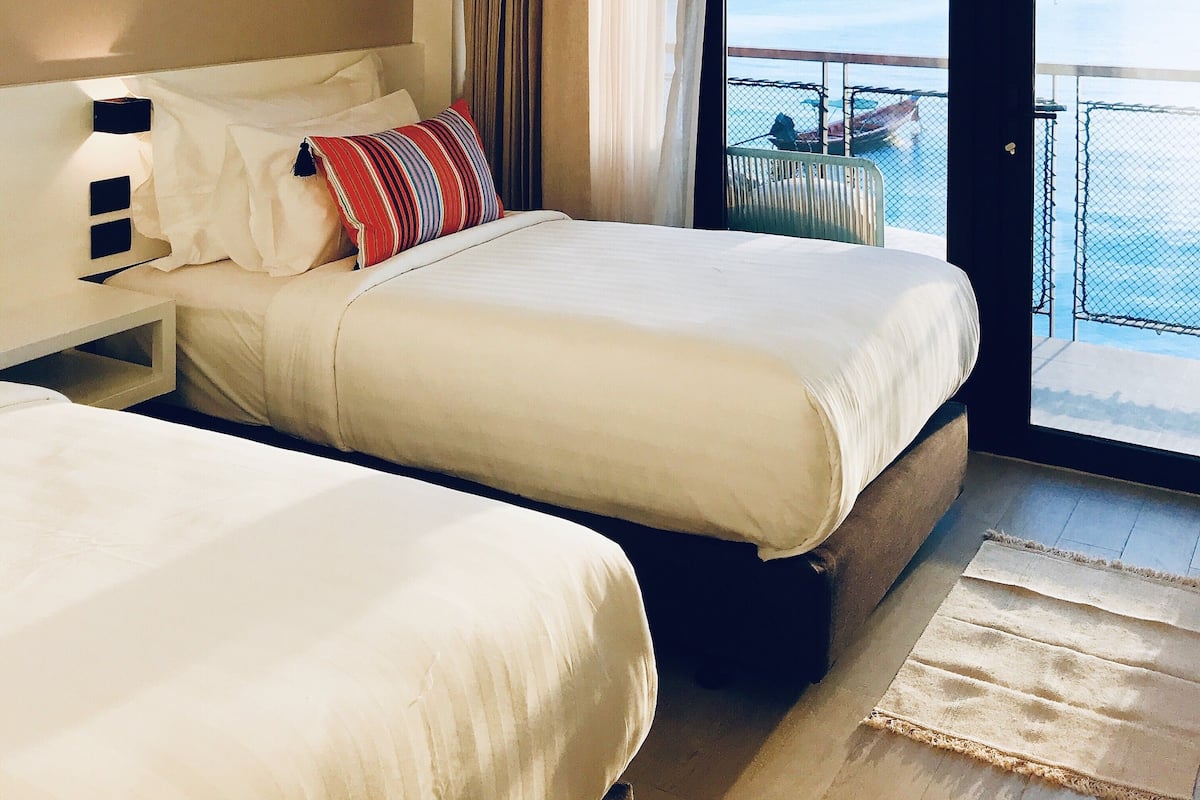 A hotel room with two beds, a nightstand, a view of the sea, and a balcony with a railing.