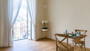 Una stanza con pavimento in legno, un tavolo da pranzo con sedie, una pianta in vaso e vista attraverso un'ampia finestra con tende bianche.