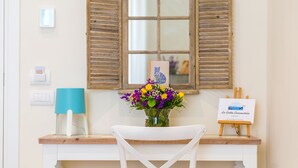 Una stanza con una scrivania in legno, una sedia bianca, un vaso con fiori, una lampada blu e un cavalletto con un dipinto.