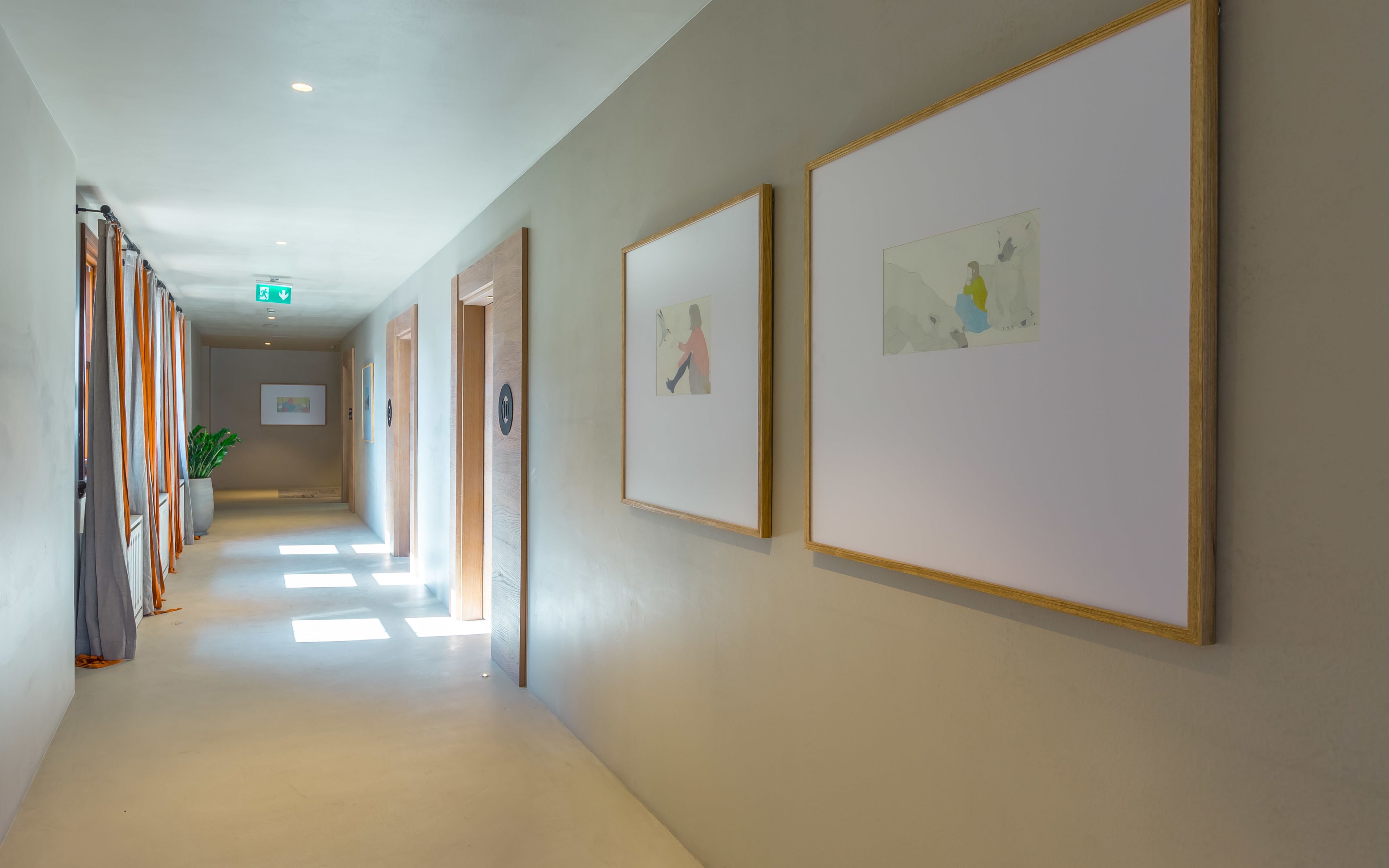 A hallway with framed artworks on the walls, a potted plant, and a sign indicating an exit.