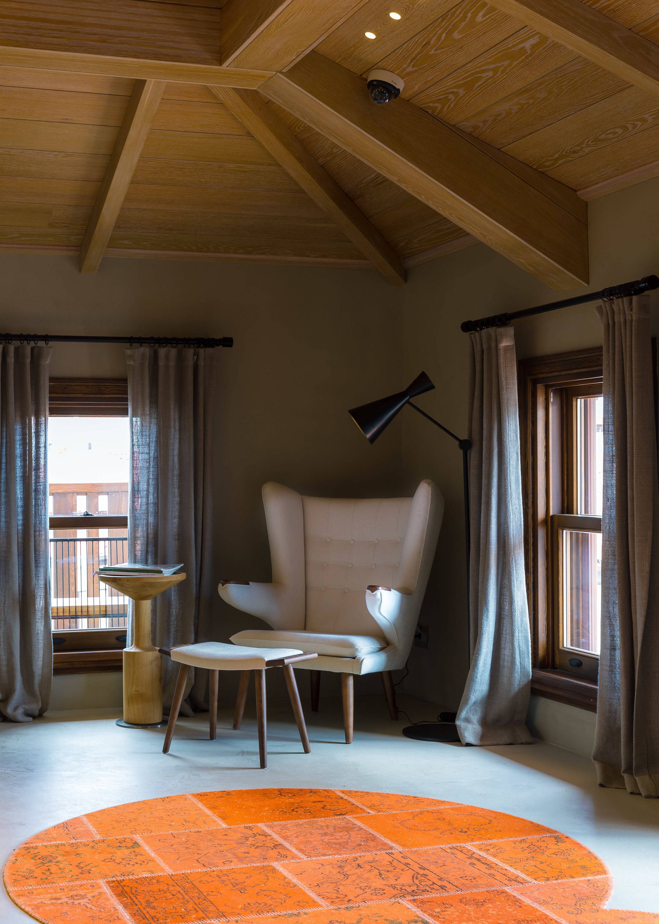 Une pièce avec un plafond haut, une grande fenêtre avec des rideaux, un fauteuil blanc, une petite table, un lampadaire et un tapis orange.