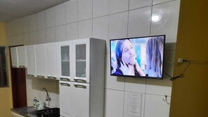 A compact kitchen with white cabinets, a built-in stove, a small dining table with two chairs, and a wall-mounted TV.
