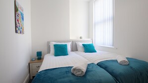 Ein Schlafzimmer mit zwei Betten, weißer Bettwäsche, türkisfarbenen Kissen, einem Nachttisch mit Lampe und einem Bild an der Wand.