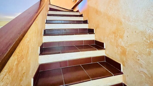 A staircase with dark tiles and wooden handrails, leading to a room with a radiator and a light switch on the wall. - Badualga Pink (San Teodoro)