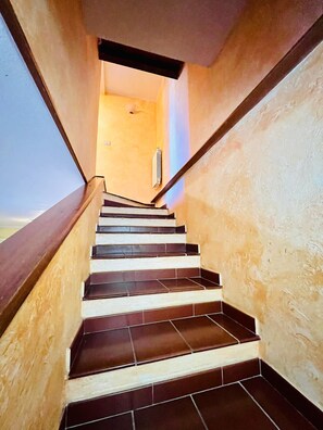 Un escalier recouvert de carreaux sombres et doté de rampes en bois, menant à une pièce où se trouvent un radiateur et un interrupteur sur le mur.