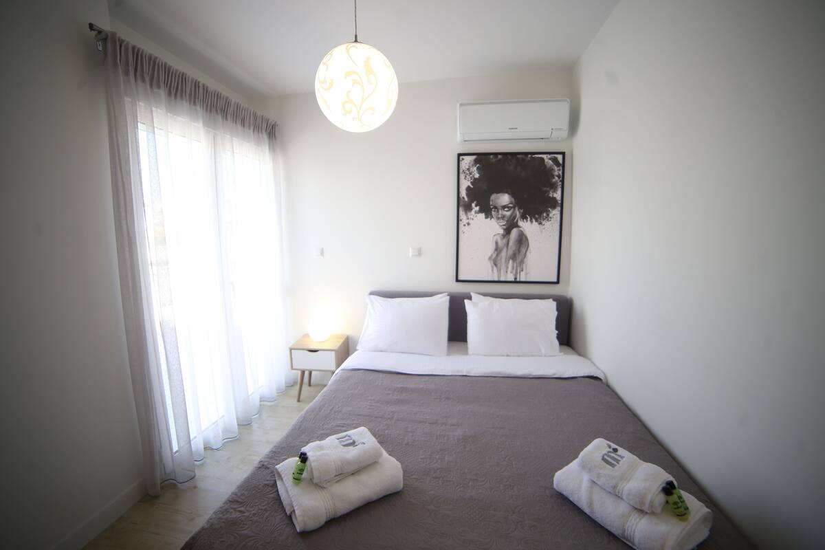 A modern hotel room with a large bed, white curtains, a hanging light fixture, and a framed artwork on the wall.