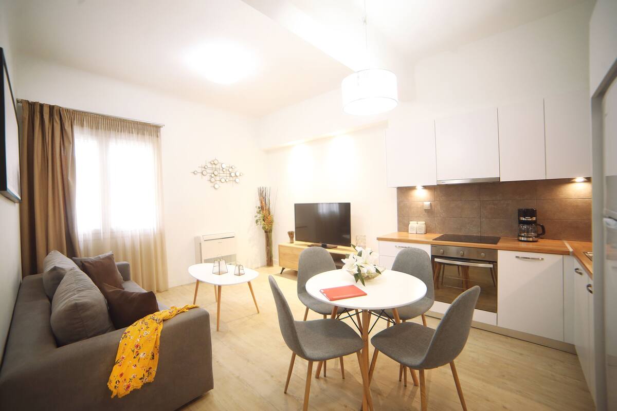 A modern living room with a sofa, a dining table with chairs, a TV, and a kitchen area with a stove and cabinets.