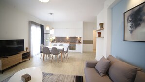 A modern living room with a flat-screen TV, a sofa, a coffee table, and a dining area with chairs and a kitchen in the background. - Athens Morum City Apartments (Vyronas)