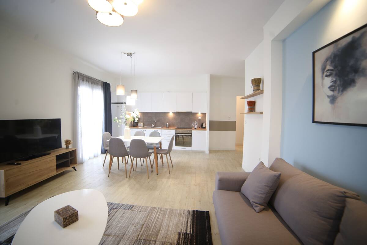 A modern living room with a flat-screen TV, a sofa, a coffee table, and a dining area with chairs and a kitchen in the background.