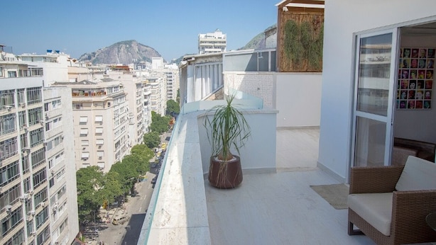A balcony with a potted plant, a wicker sofa, and a glass door leading to a room with a view of a cityscape and mountains.