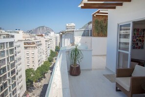 En balkon med en potteplante, en fletsofa og en glasdør, der fører til et rum med udsigt over by og bjerglandskab.