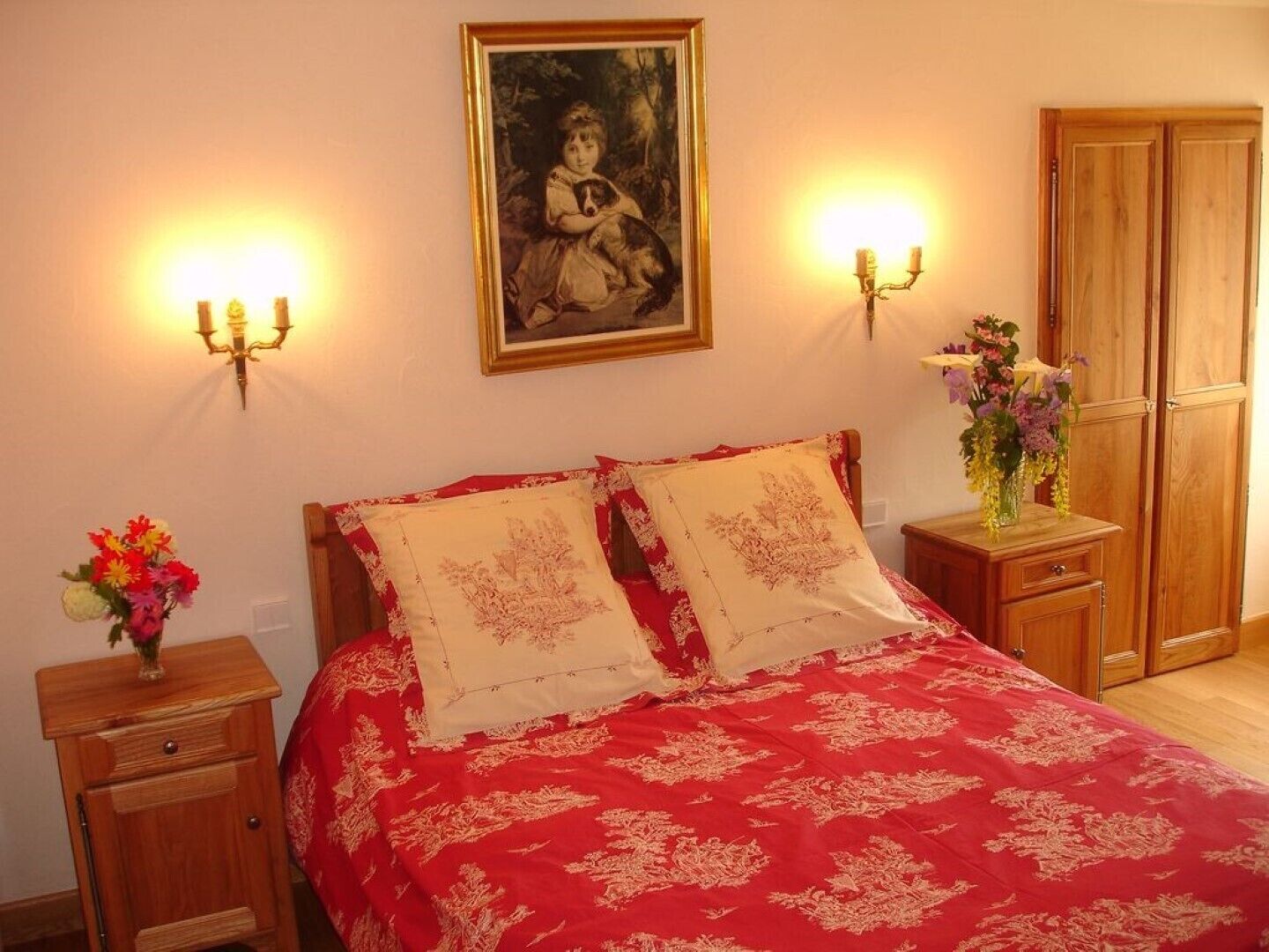 A bedroom with a red bedspread, two matching pillows, a nightstand with a vase of flowers, a wooden wardrobe, and a wall-mounted painting.