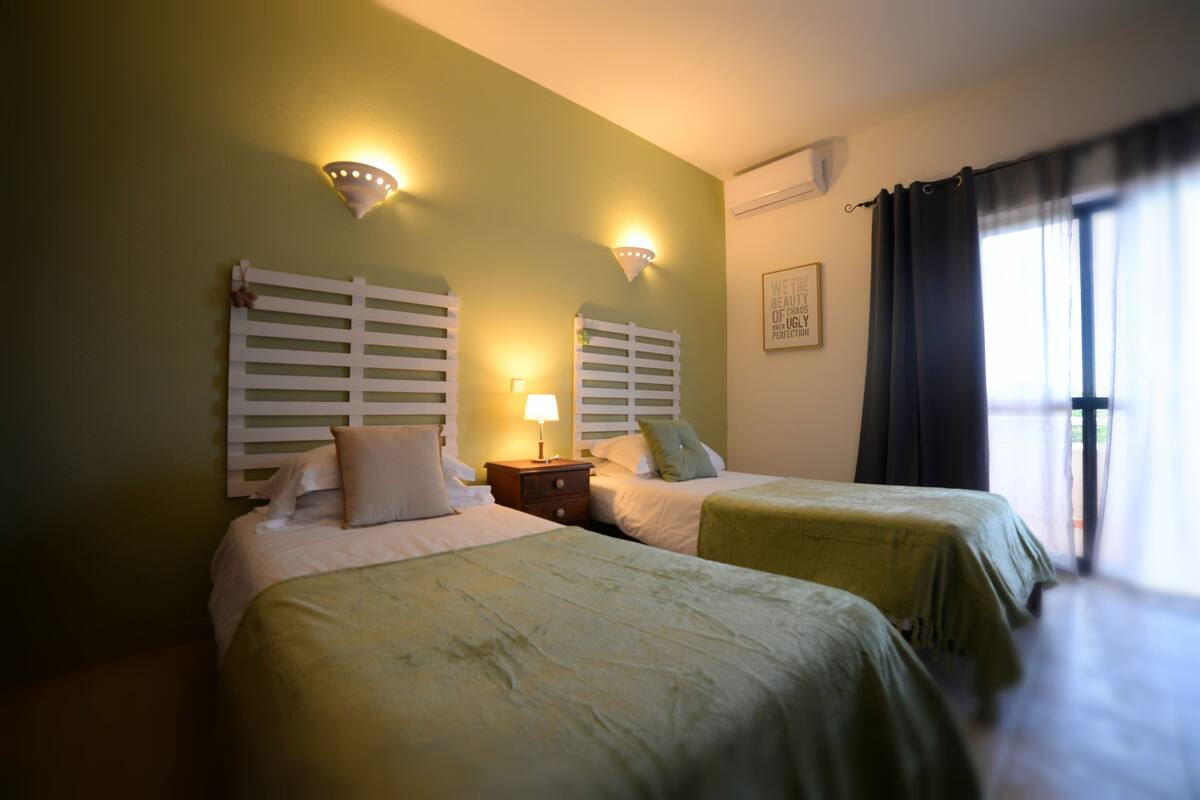 A hotel room with two beds, a nightstand, wall-mounted lights, and a window with curtains.
