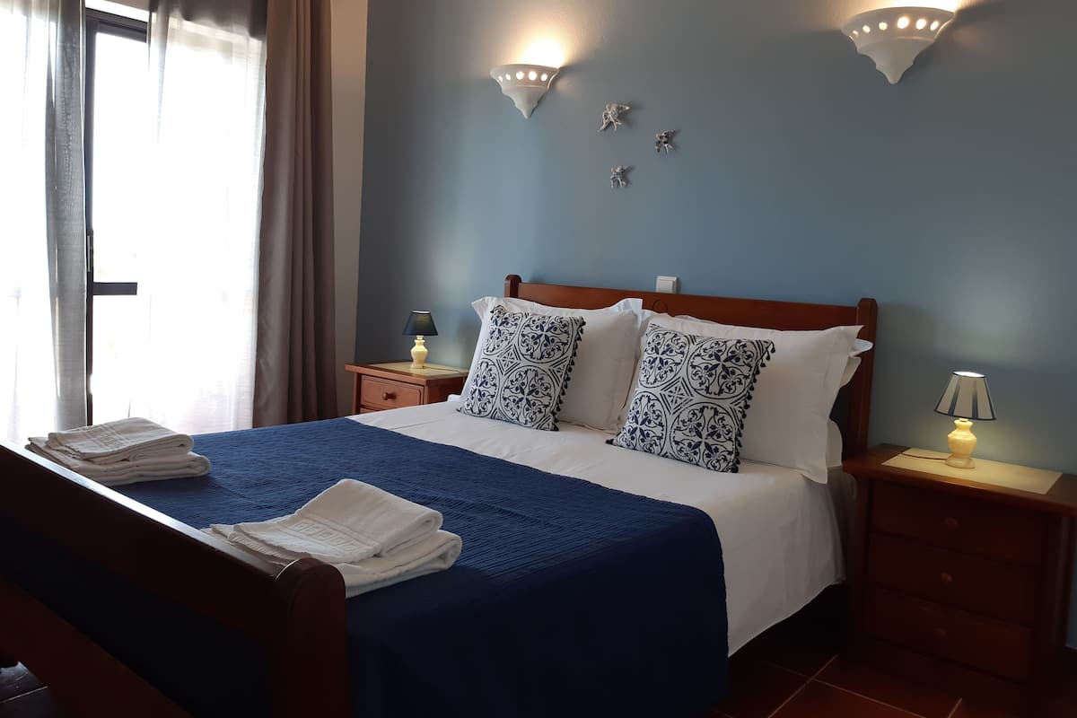 A neatly made bed with a blue bedspread and white pillows, two bedside lamps, a wooden nightstand, and a window with curtains.