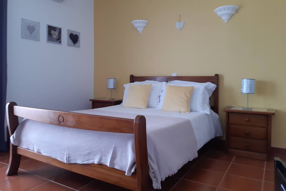 A single bed with a wooden frame and white bedding, two bedside tables with lamps, a wall with three heart-shaped decorations, and a tiled floor.