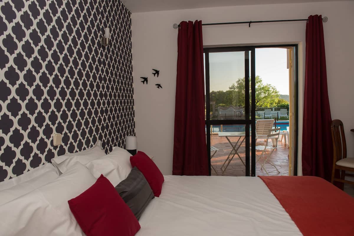 A hotel room with a bed, red curtains, a view of an outdoor pool area, and a patterned wall.