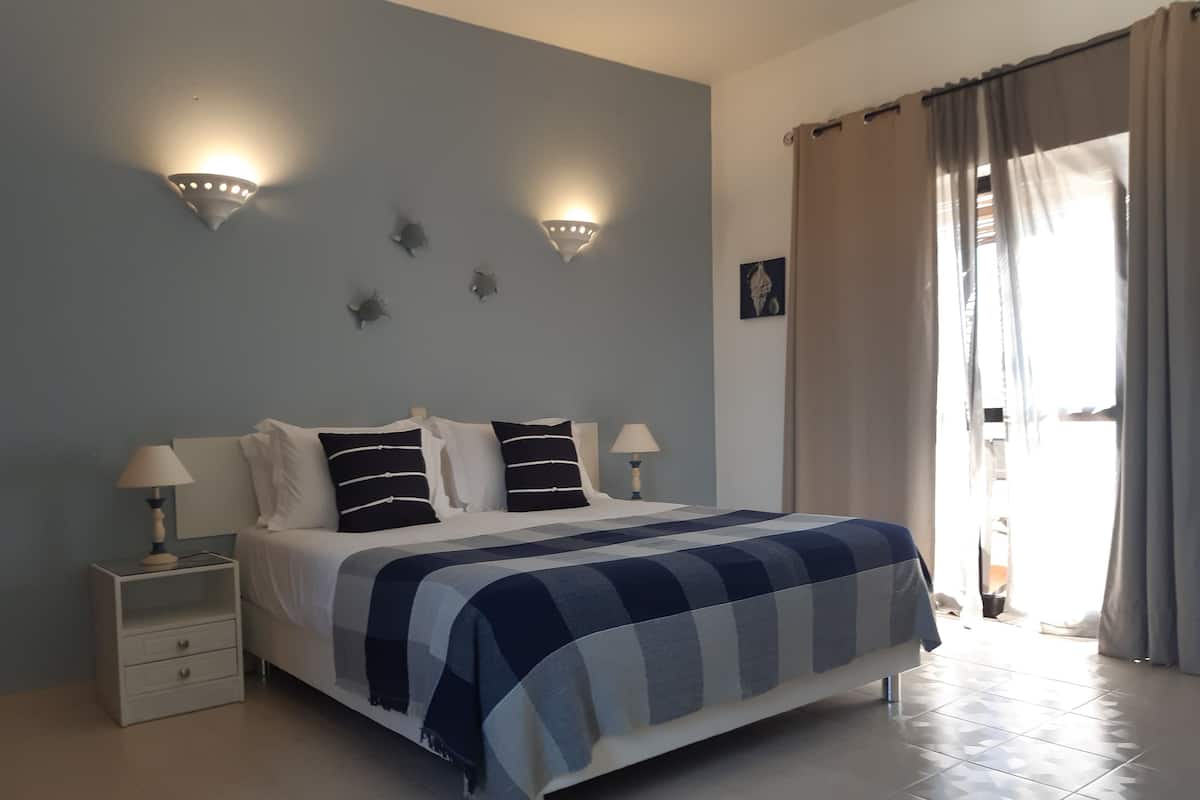 A bedroom with a bed, two bedside tables, a wall-mounted light fixture, a window with curtains, and a small wall decoration.