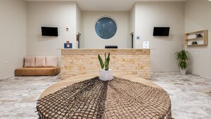 A reception area with a stone wall, a round table with a woven centerpiece, a sofa, two TVs, and a large window. - Sisi Breeze Hotel (Agios Nikolaos)