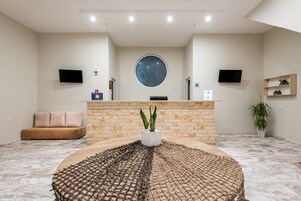 Un área de recepción con pared de piedra, una mesa redonda con un centro de mesa tejido, un sofá, dos televisores y un amplio ventanal.