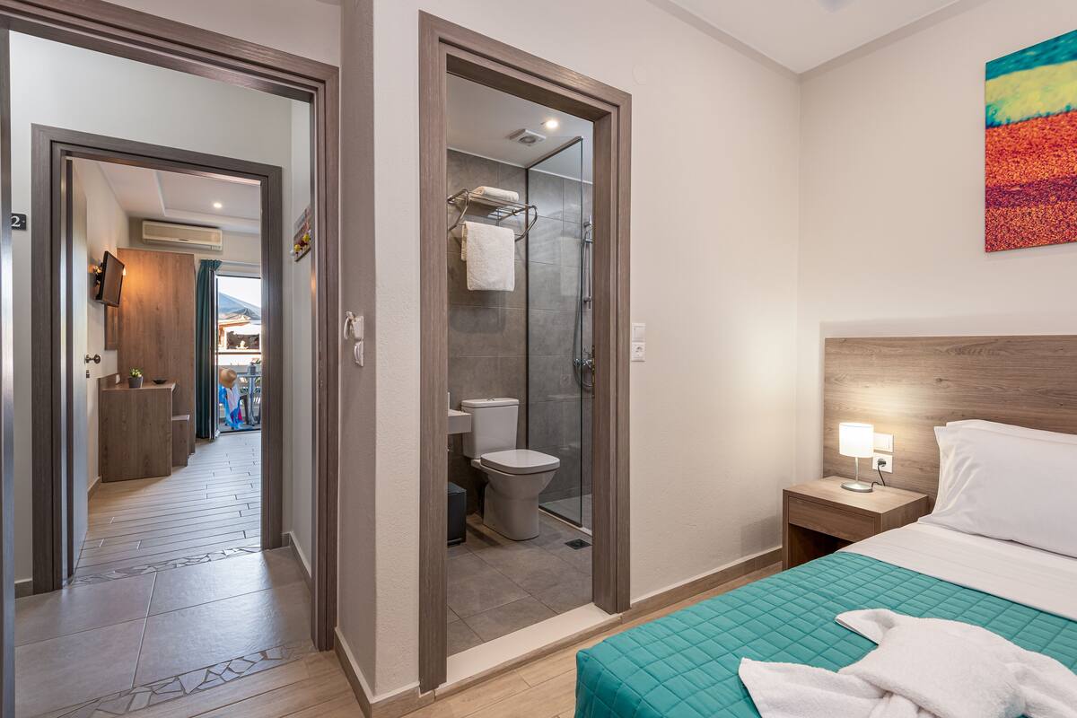A hotel room with a bed, a nightstand, a bathroom with a shower, and a hallway leading to other rooms.