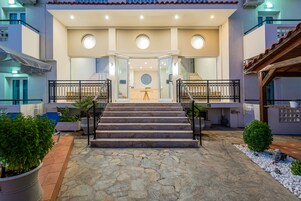 La entrada de un hotel con un camino empedrado que conduce a un conjunto de escalones, una puerta de cristal y una pequeña zona ajardinada con plantas en macetas.