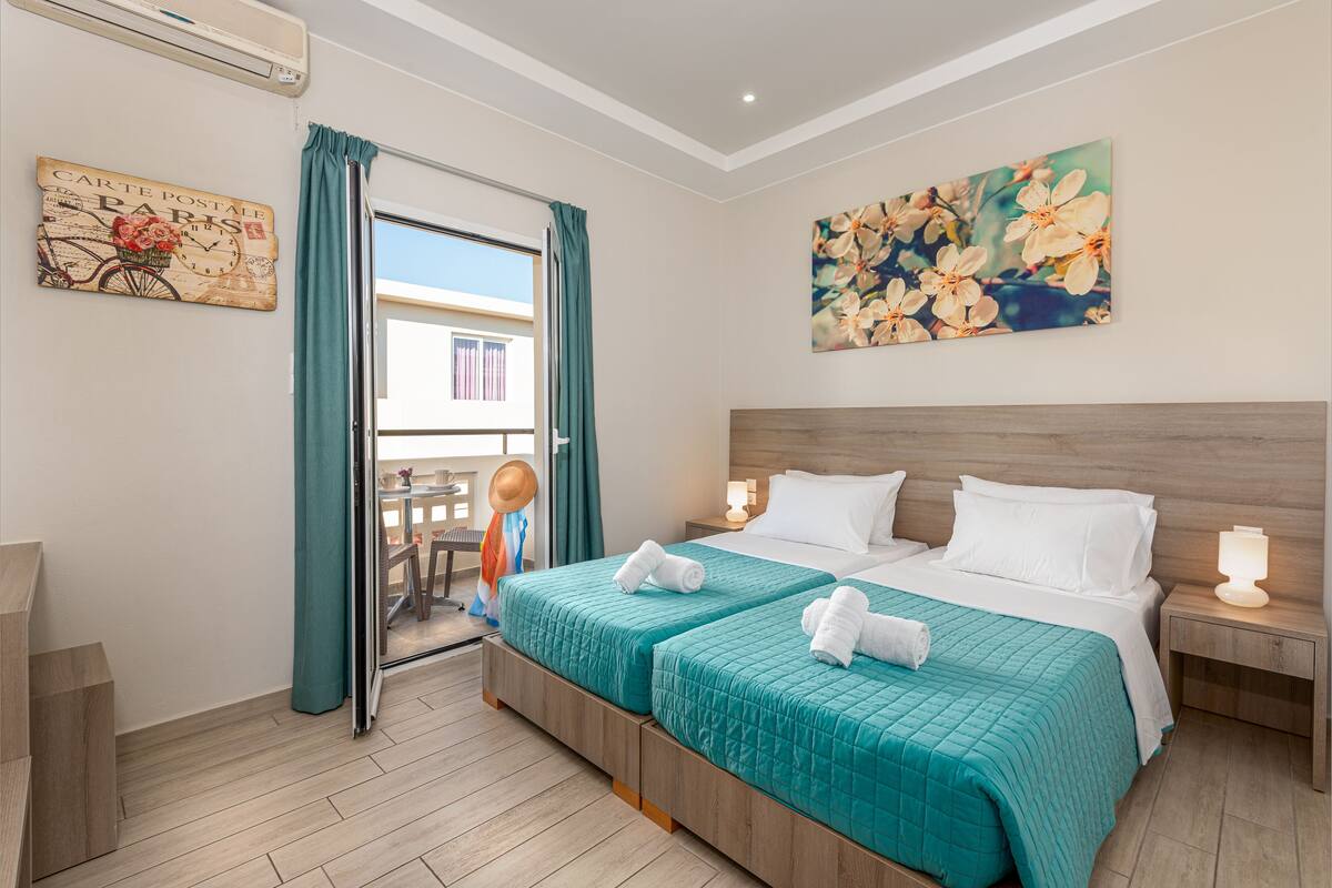 A hotel room with two beds, teal bedding, a wooden headboard, a painting of flowers, and a view of a balcony with a chair and a hat.