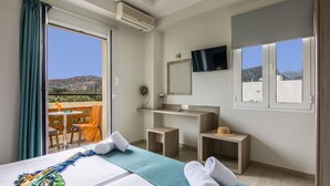 A hotel room with a bed, a small desk, a mirror, a TV, and a balcony with a view of the hills. - Sisi Breeze Hotel (Agios Nikolaos)