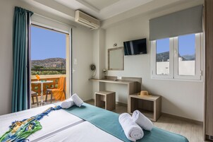 Habitación de hotel con cama, un escritorio pequeño, espejo, televisor y balcón con vistas a las colinas.
