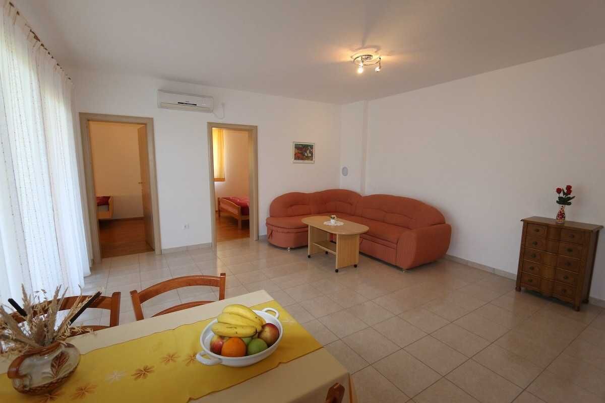 A living room with a sofa, a dining table with fruit, a small table with a vase, and a cabinet.
