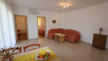 A living room with a sofa, a dining table with fruit, a small table with a vase, and a cabinet.