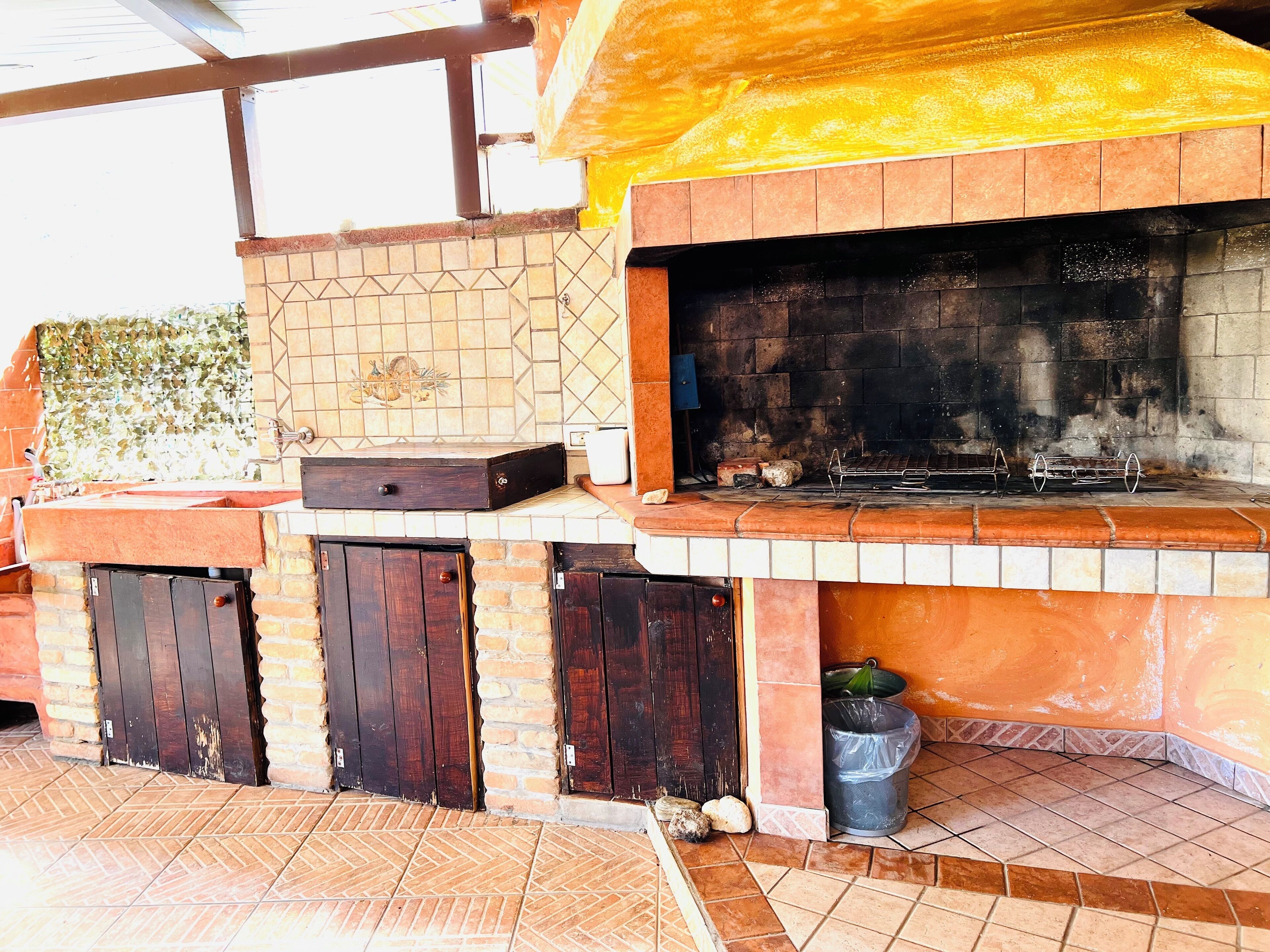 An outdoor kitchen with a tiled backsplash, a grill, and a trash can.